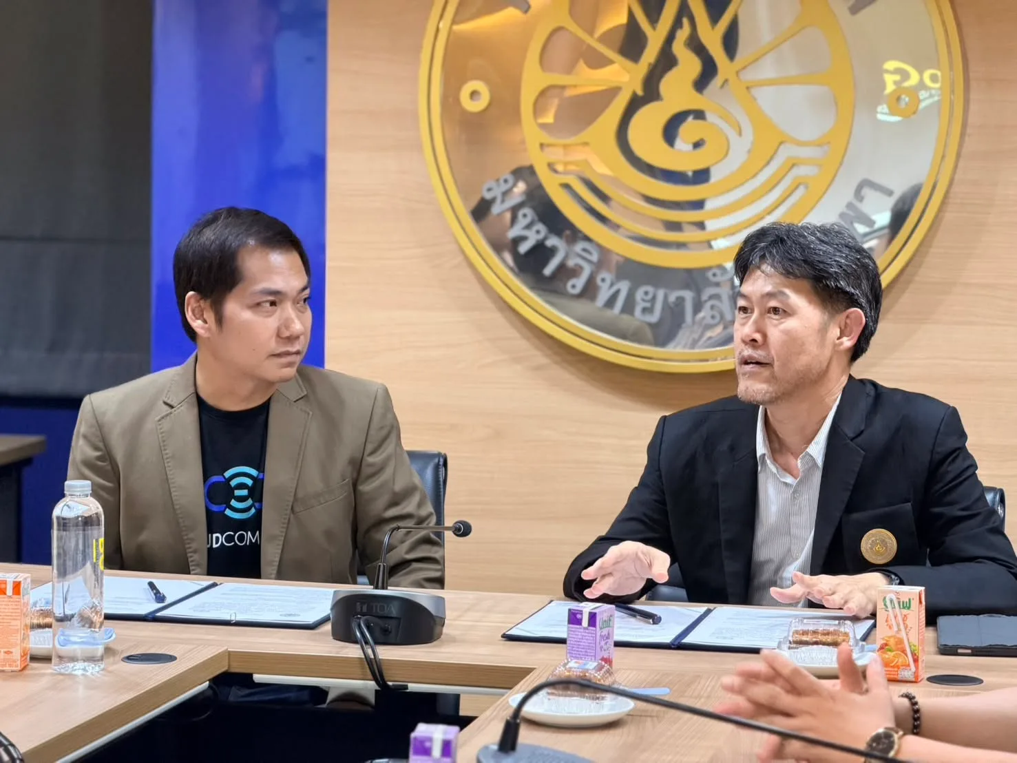 A university representative speaks while a CloudCommerce representative listens, in front of the Burapha University emblem